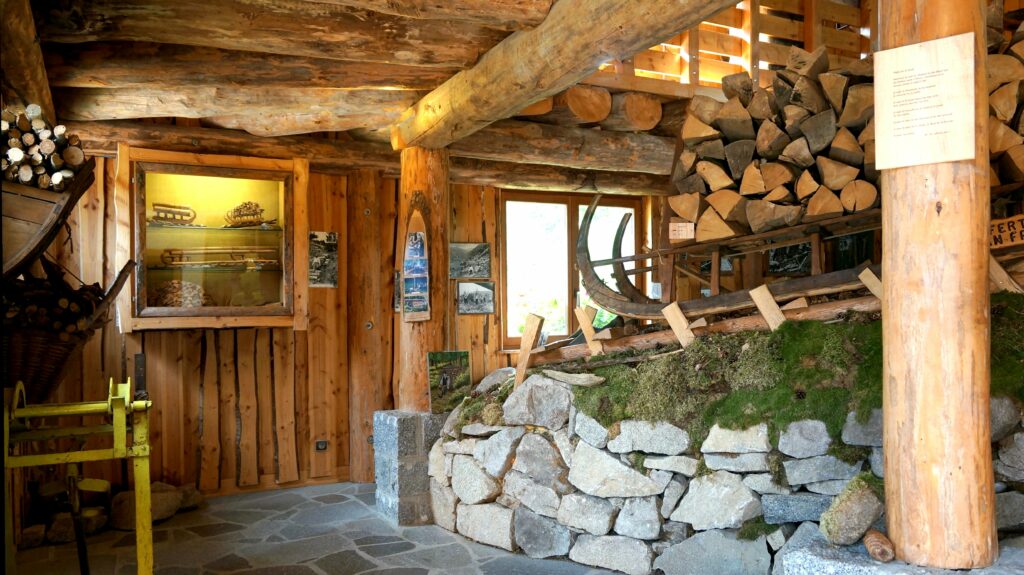 Rustic log cabin interior with large wooden beams, stacked firewood, and stone wall, displaying historical photos and tools in a museum-like space.