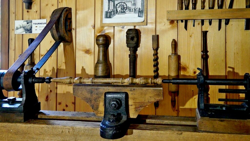 Antique wood lathe on a wooden shop bench with turning chisels on a rack and a crank drive in a bright workshop wall paneling