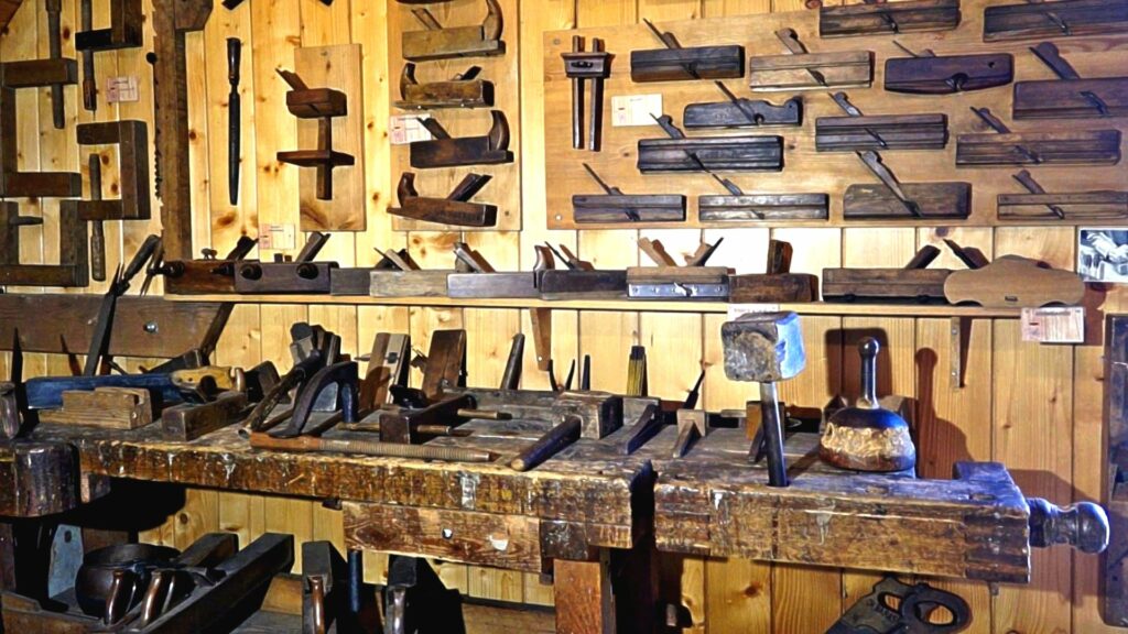 Woodworking workshop with dozens of hand planes mounted on pegboard and a heavy workbench cluttered with chisels and hammers.